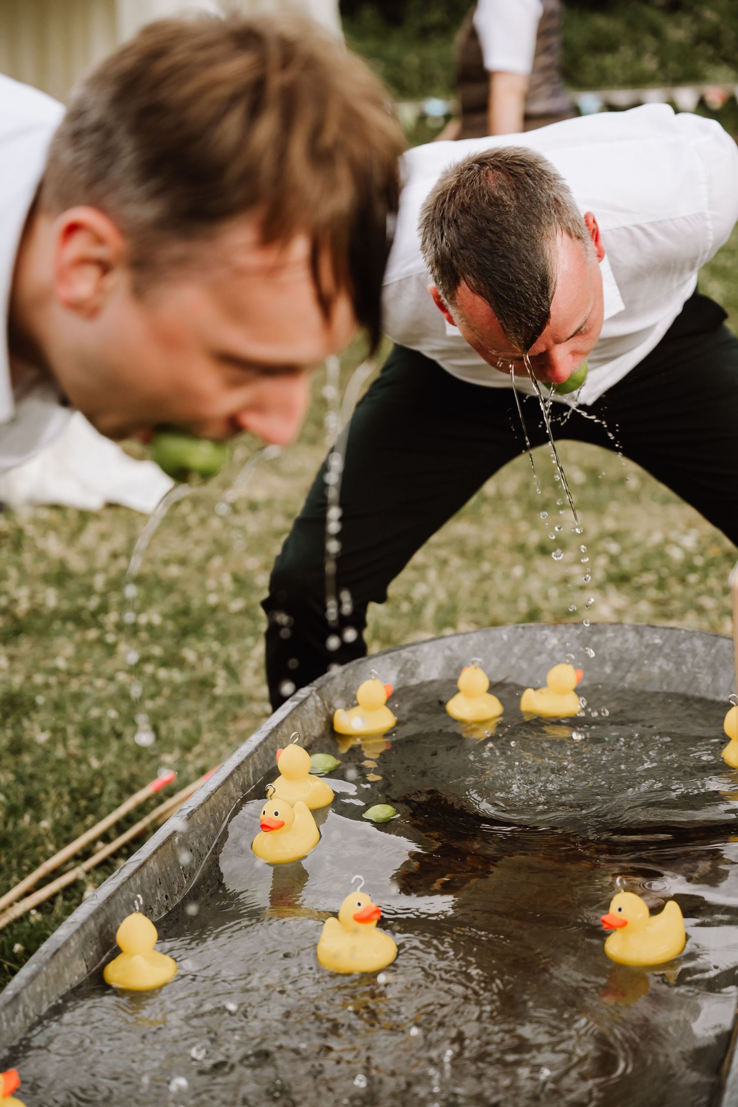 Two men bobbing for rubber ducks in a metal tub outdoors at a garden party game.