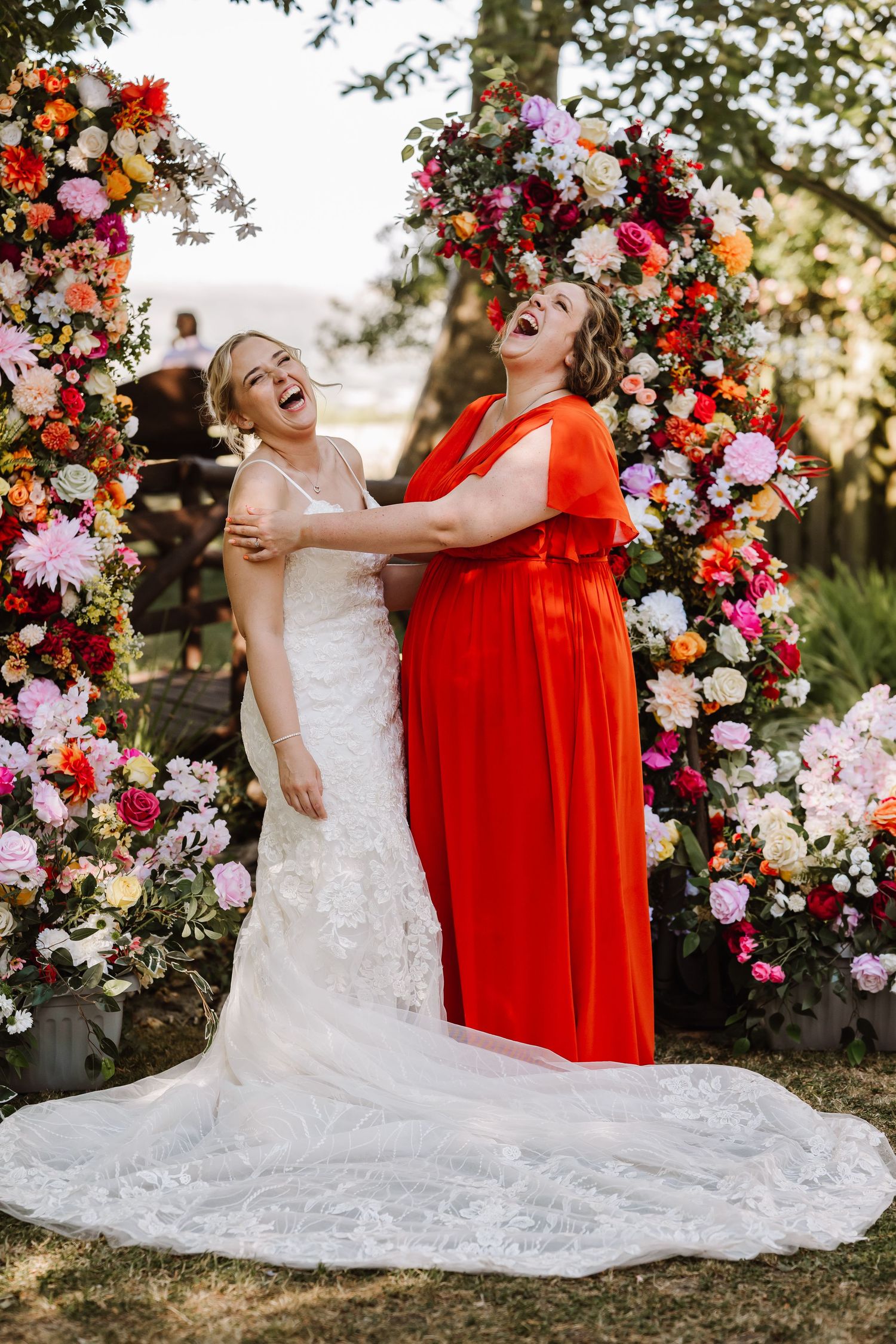 Two brides laughing joyfully in front of a colorful floral arch, one in white gown and one in red dress.