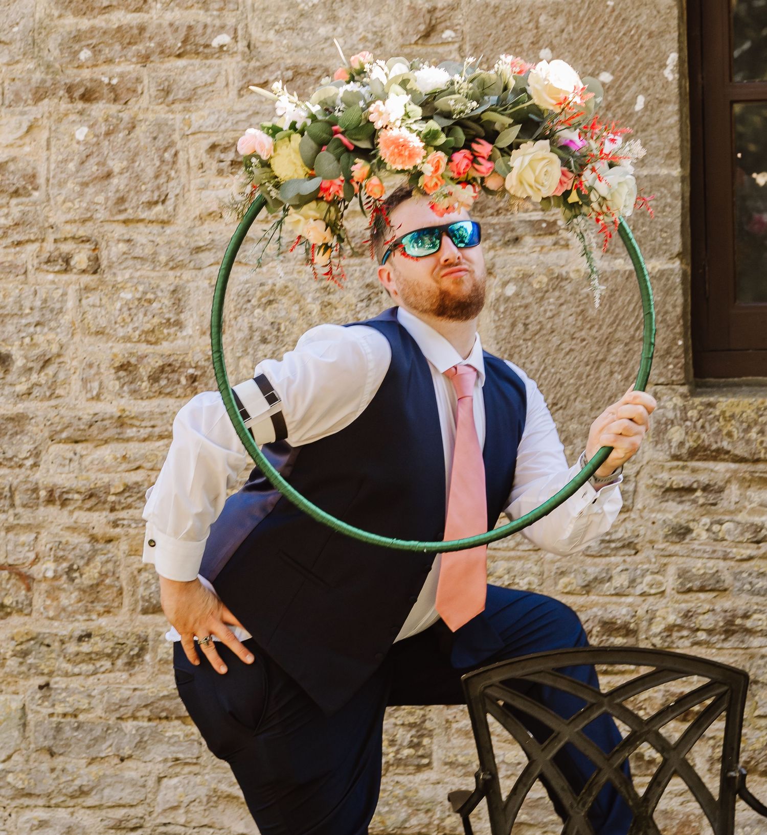 Groom in navy vest and pink tie poses playfully with a floral hoop at a rustic stone wedding venue.