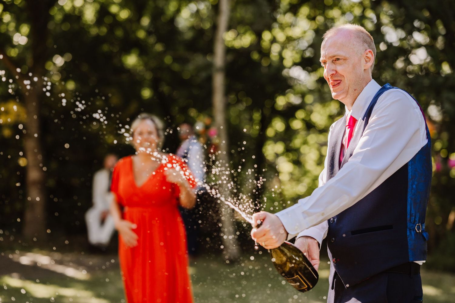 Groom pops champagne bottle outdoors at wedding celebration, with guests in background.