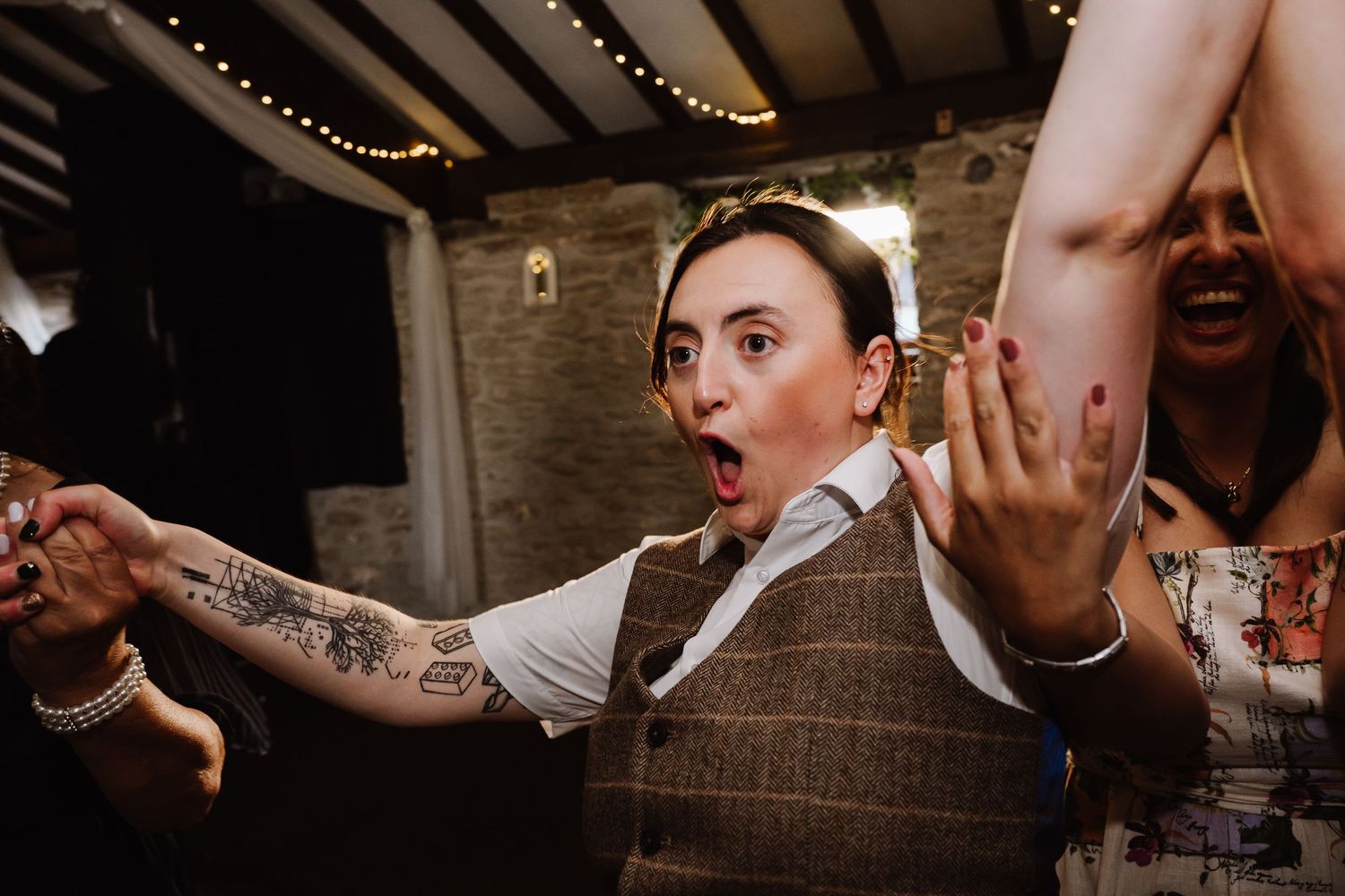 Woman with tattoos in a tweed vest dancing expressively at a wedding reception in a rustic venue.