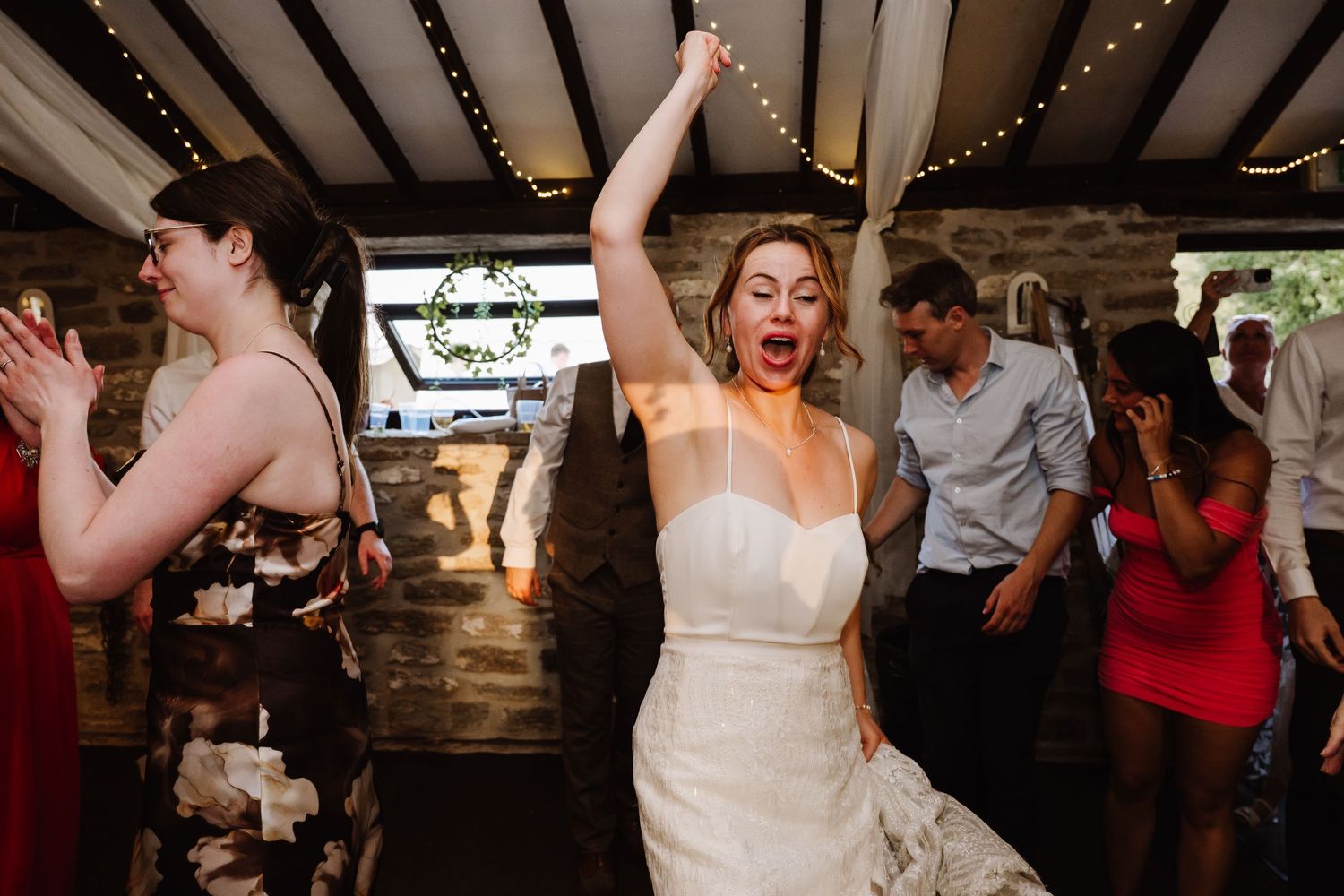 Bride in white gown dancing joyfully with arm raised at wedding reception in rustic stone venue.