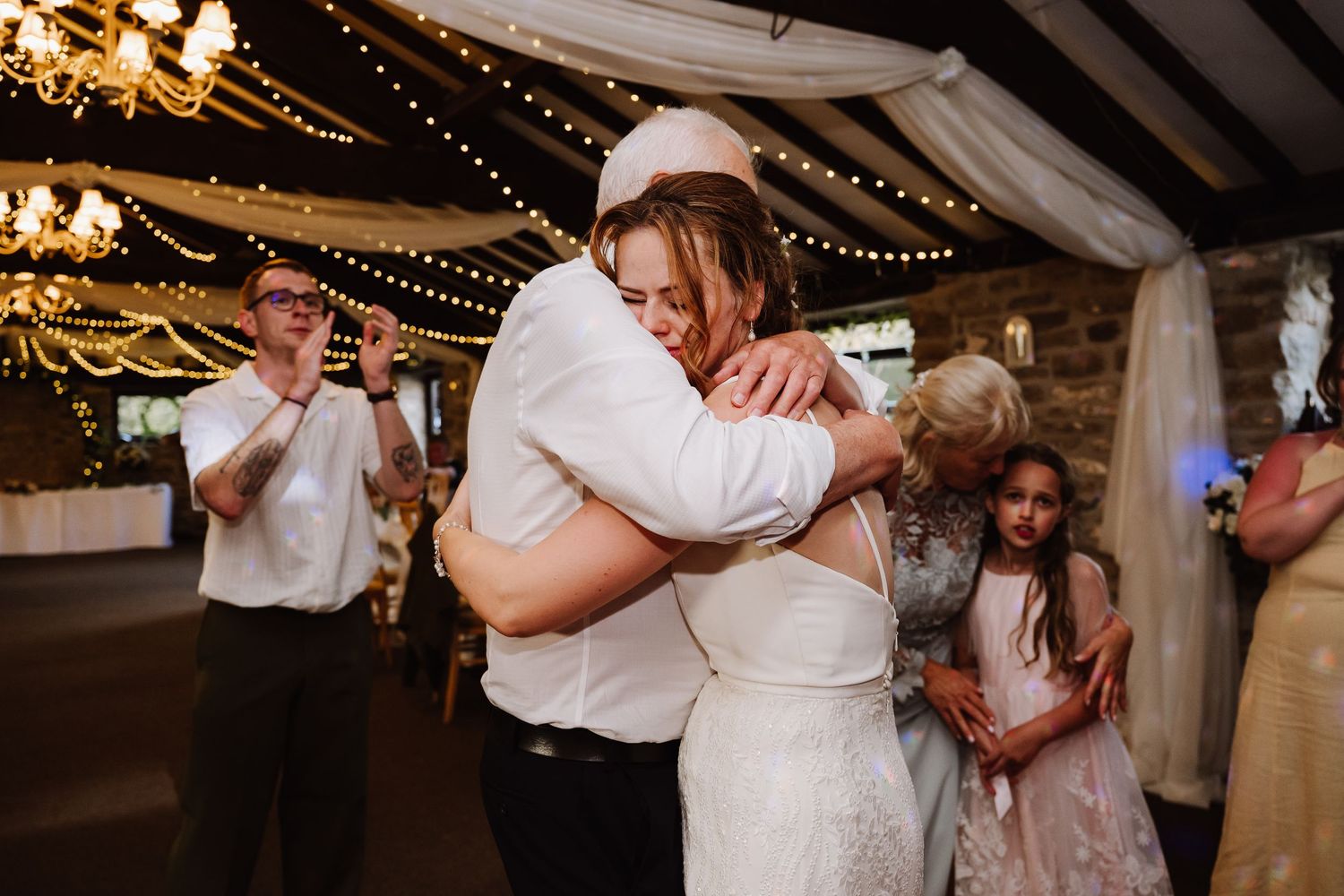 Bride and groom share a warm embrace during wedding reception in rustic venue with string lights.