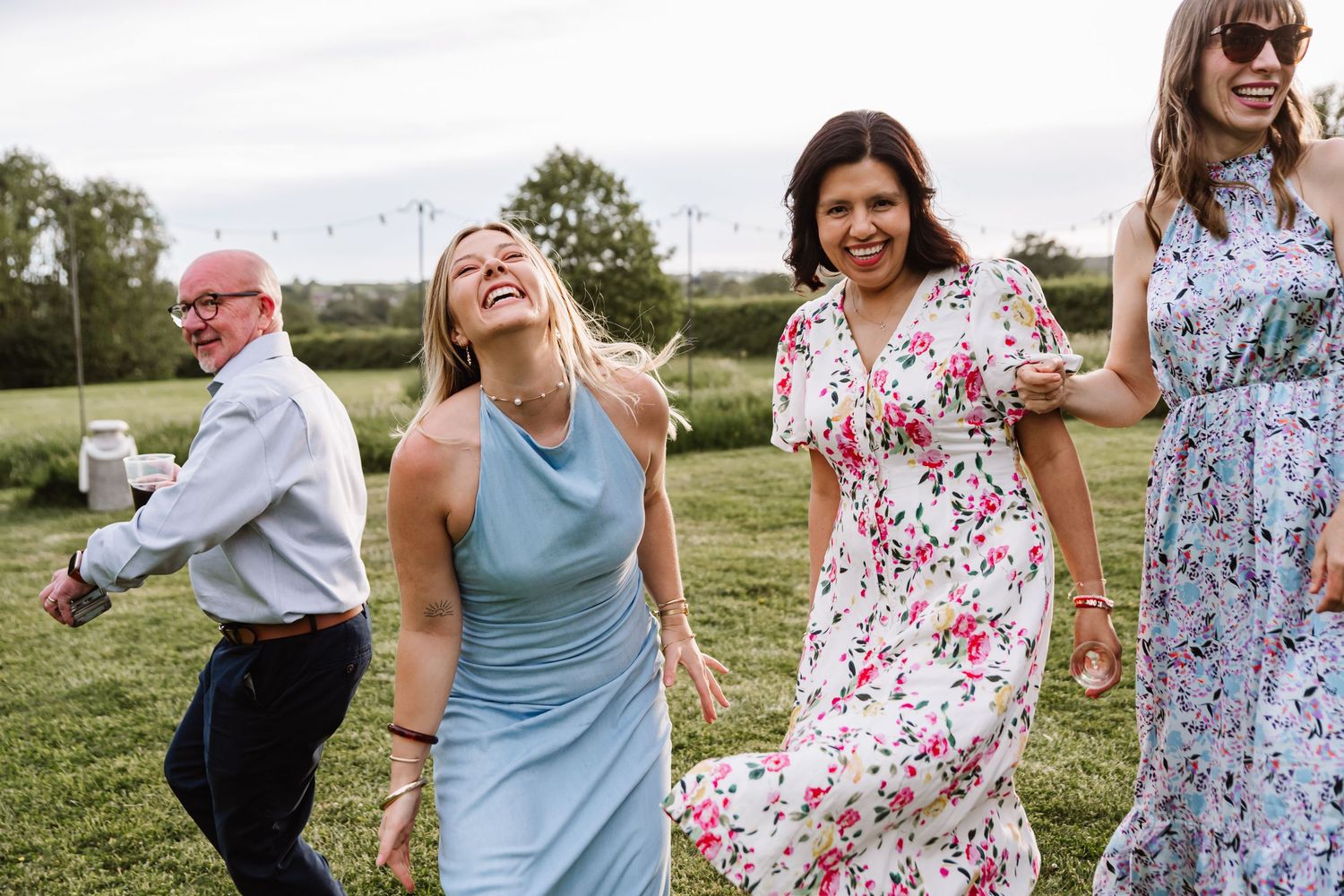 Four guests laughing and walking outdoors at a wedding celebration in a green field, wearing elegant dresses and formal attire.