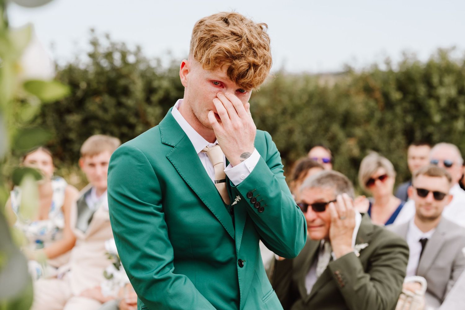 Emotional groom in green suit wiping tears at outdoor wedding ceremony with guests in background.