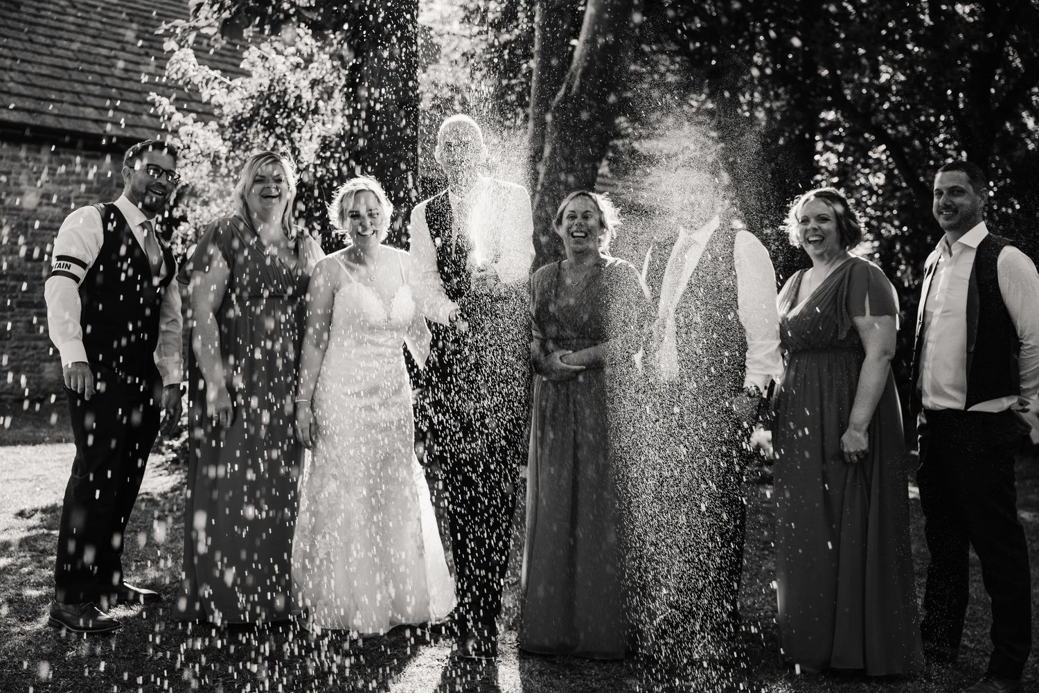 Black and white wedding party photo with champagne spray celebration outdoors near rustic barn building.