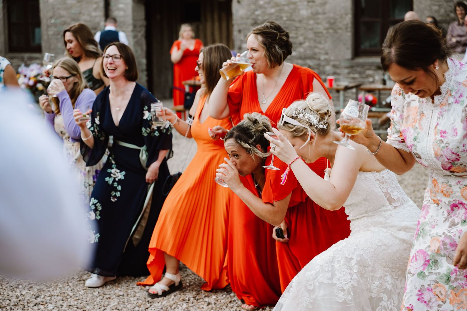 Bride and bridesmaids in colorful dresses laugh and celebrate outdoors at a wedding reception with drinks.
