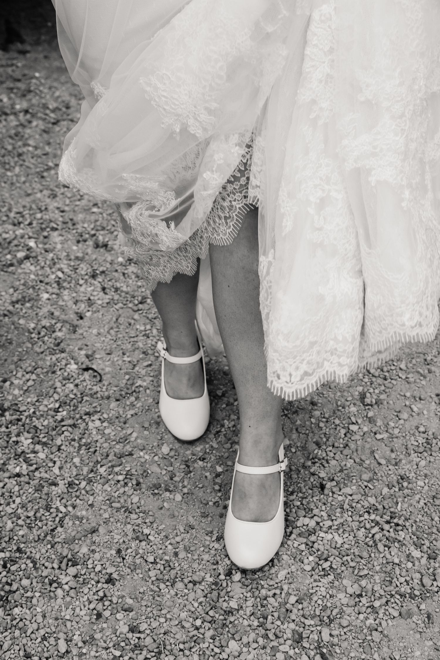 Black and white photo of a bride lifting her lace wedding dress to reveal white Mary Jane heels on a gravel path.