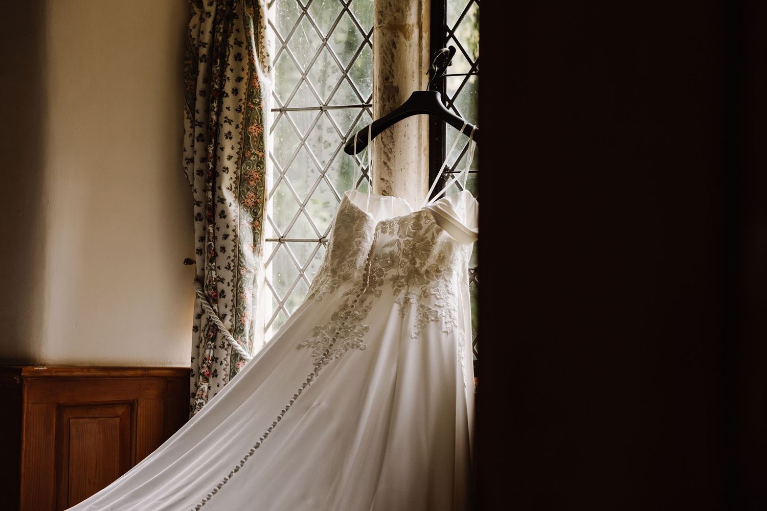 Elegant embellished wedding dress hanging on hanger in front of leaded church window at Elmhay, Orchardleigh estate, Frome