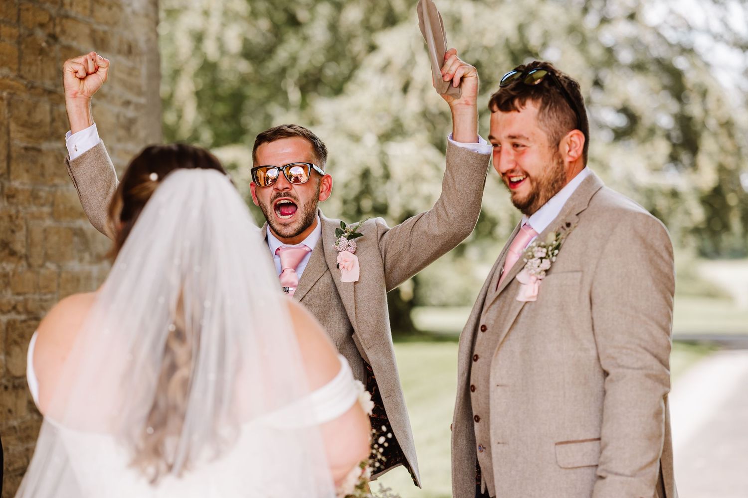 Two groomsmen in tan suits celebrating with raised arms as bride arrives at outdoor ceremony at Elmhay, Orchardleigh estate, Frome