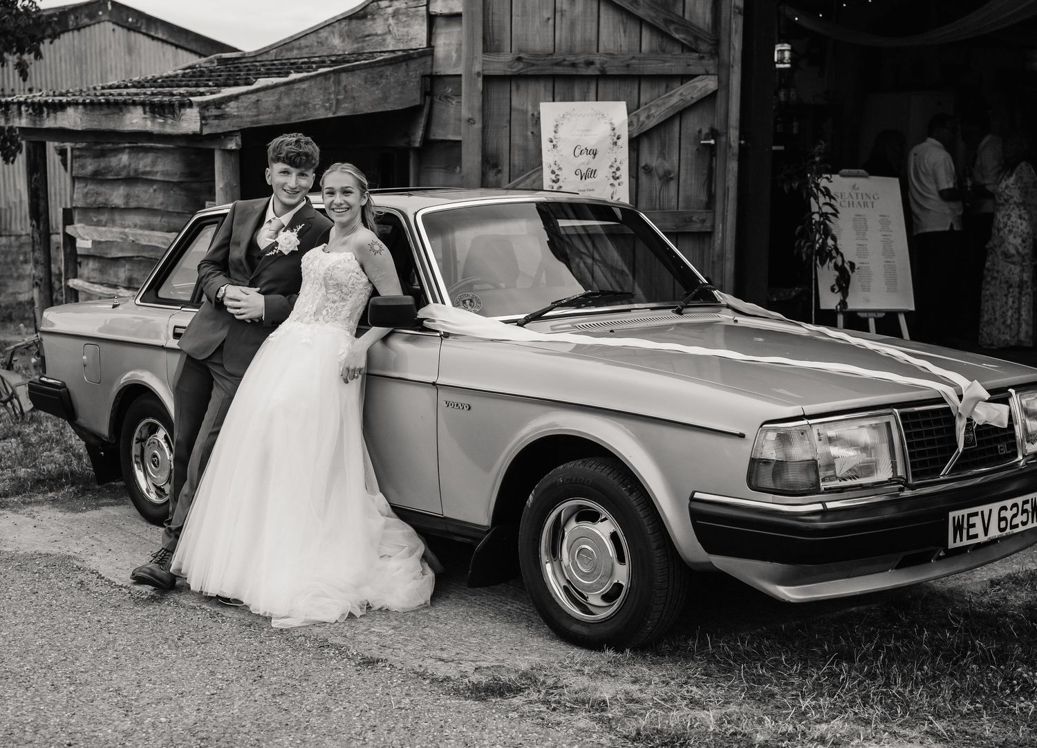 Bride and groom posing beside a classic vintage car outside a rustic barn venue in black and white wedding photo.