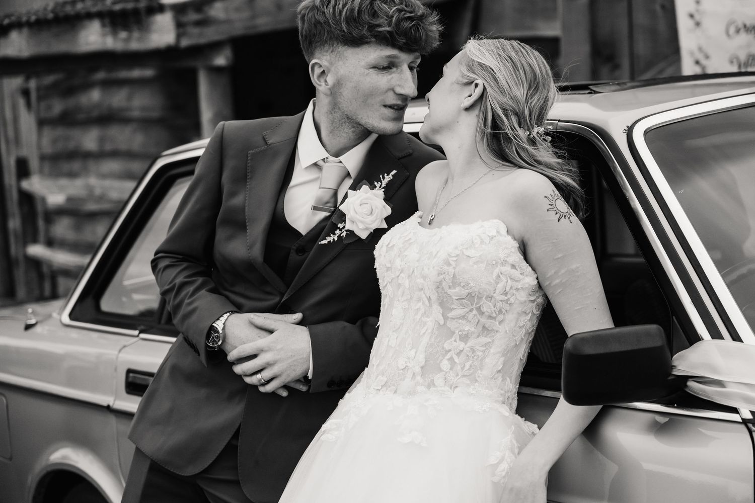 Black and white wedding photo of bride and groom sharing a romantic moment leaning against a vintage car.