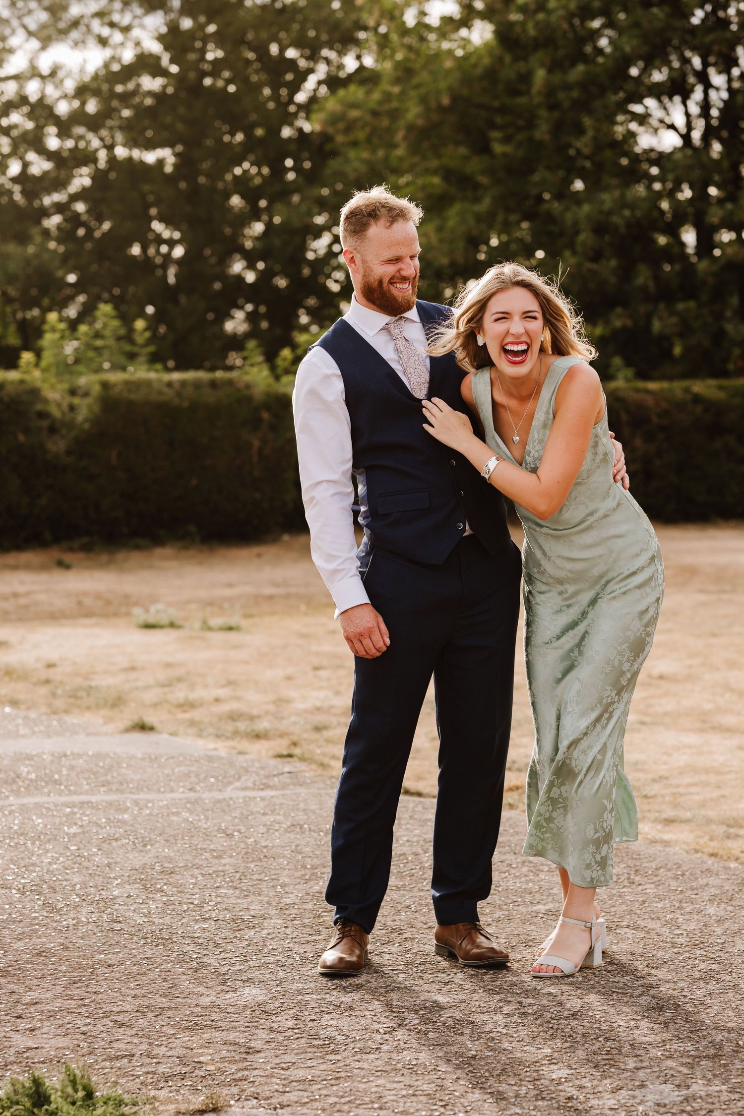 A laughing couple in wedding attire poses outdoors, the groom in navy suit and bride in sage green dress.