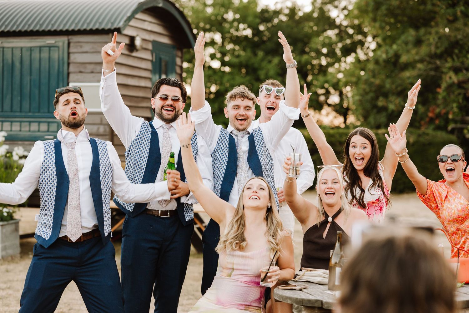 Joyful wedding party celebrating outdoors, groomsmen in blue vests raising hands with bride in pink dress.