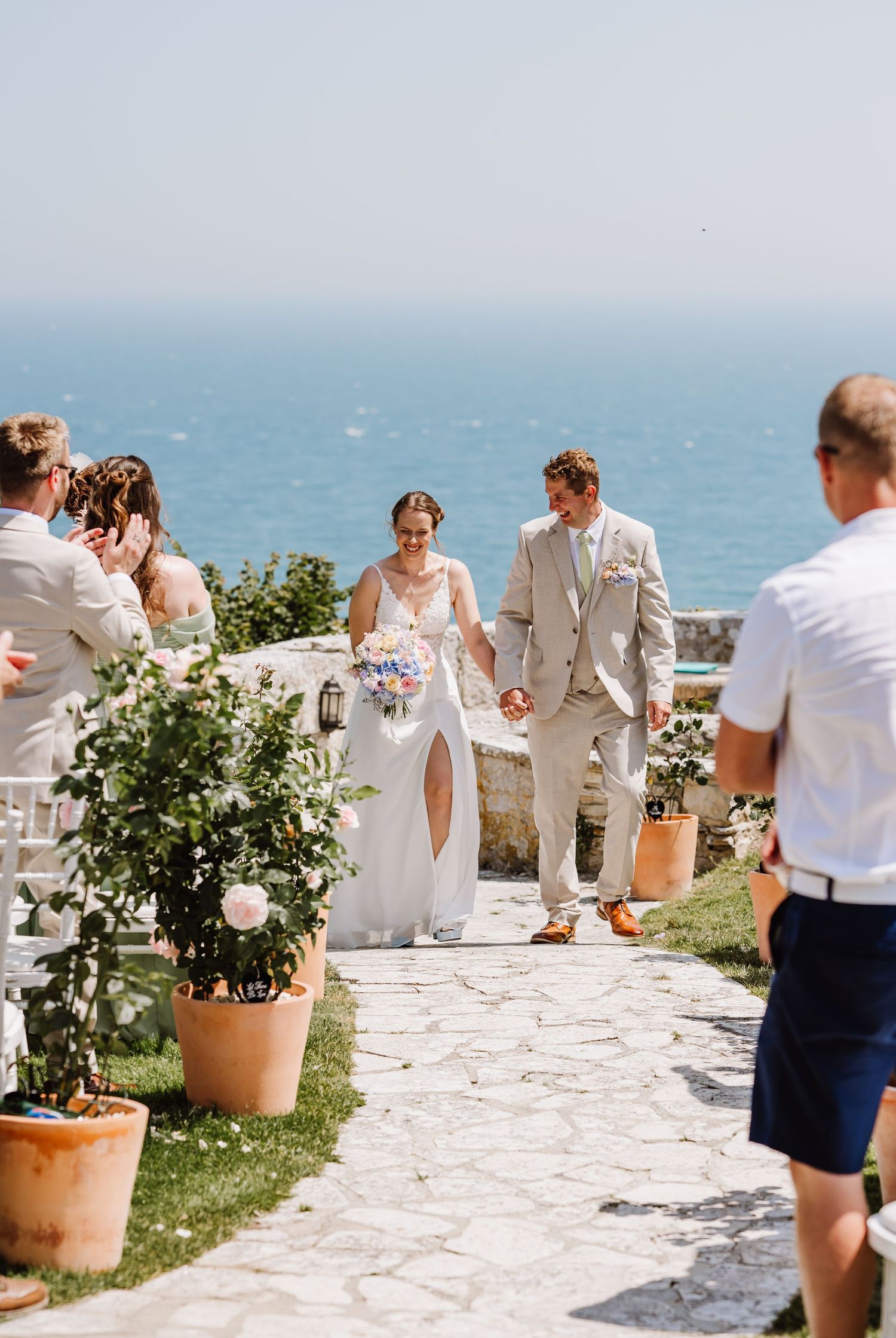 Bride and groom walk down outdoor aisle with ocean backdrop at sunny coastal wedding ceremony at Pennsylvania Castle, Portland Dorset