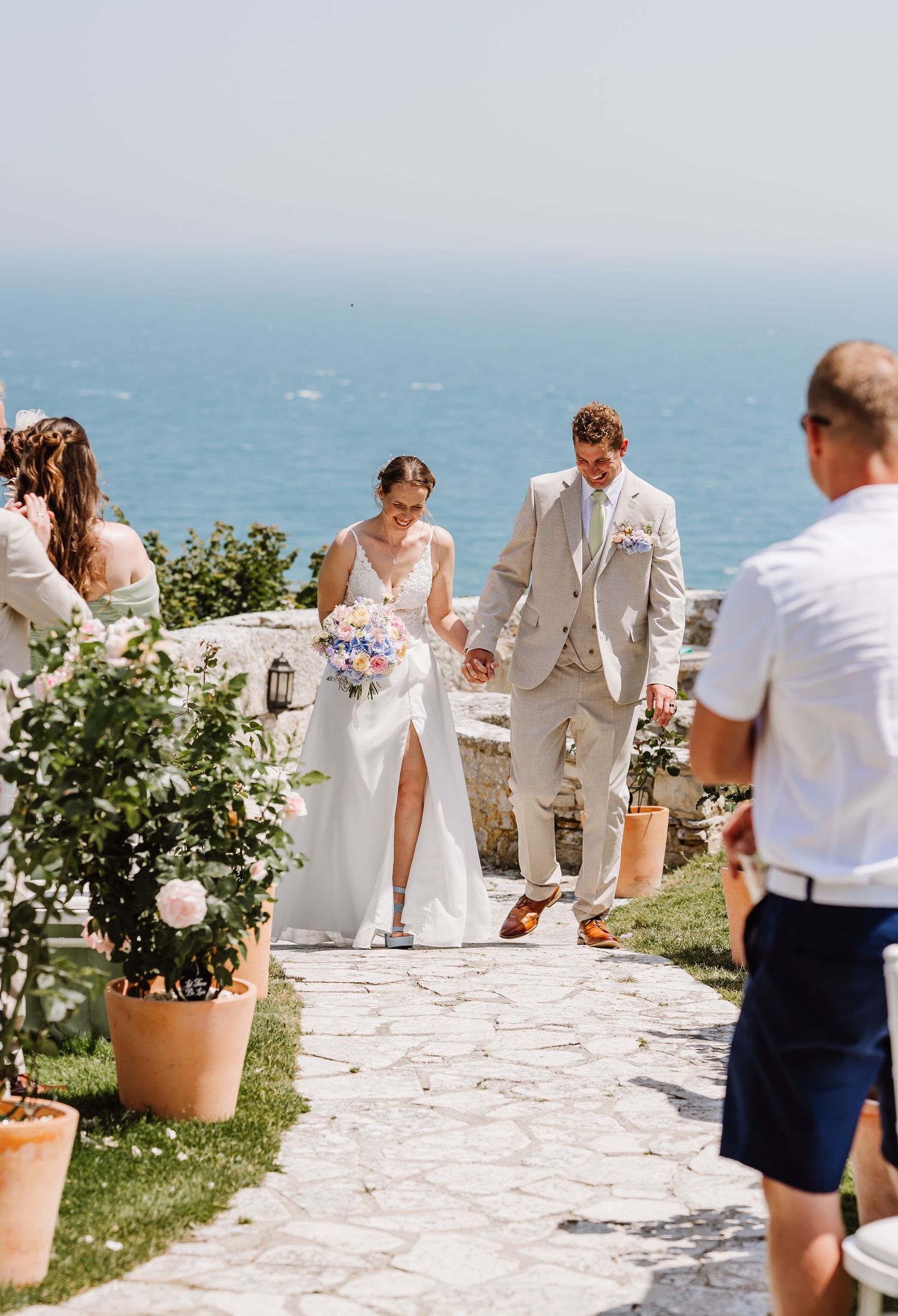Newlyweds hold hands walking down stone path aisle with sea view at Pennsylvania Castle, Portland Dorset
