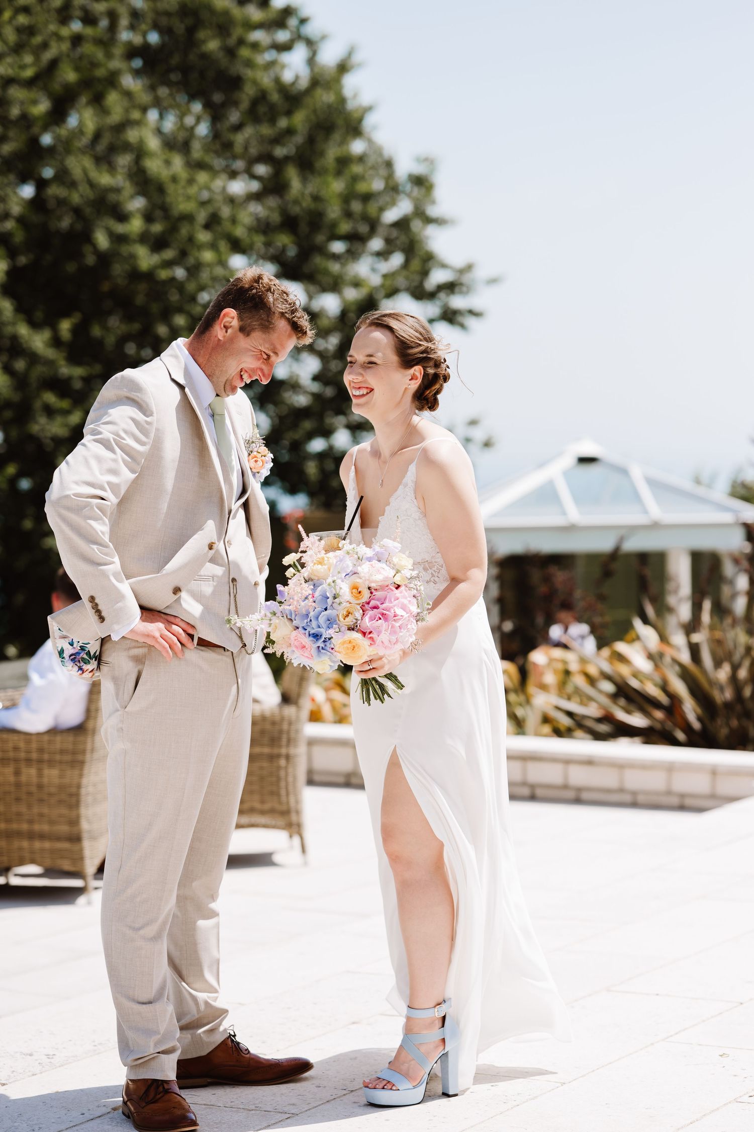 Smiling bride holding colorful bouquet laughs with groom in beige suit at outdoor wedding at Pennsylvania Castle Portland, Dorset