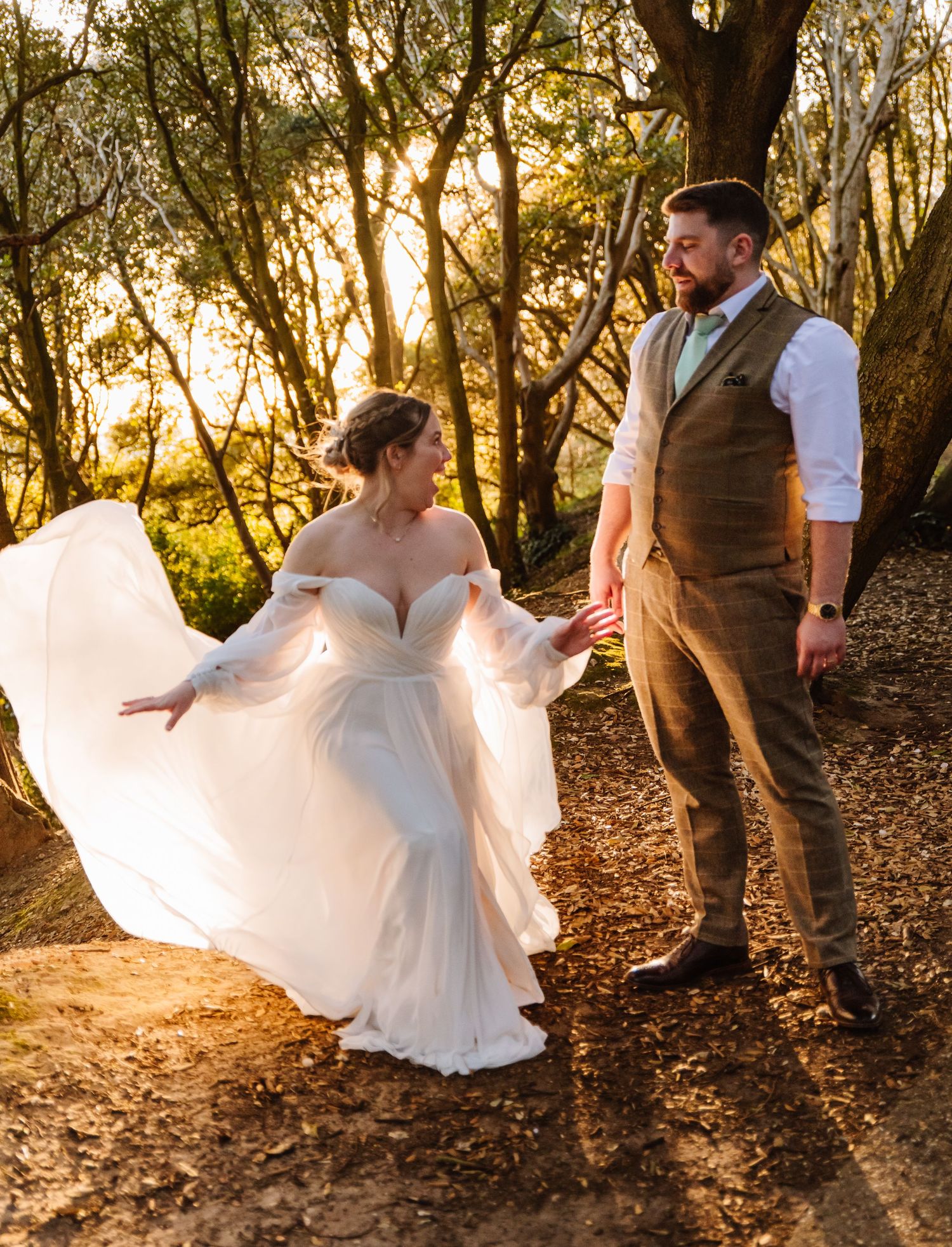 Bride in flowing white gown holds groom's hand in sunlit forest, golden hour light illuminating their romantic wedding portrait.