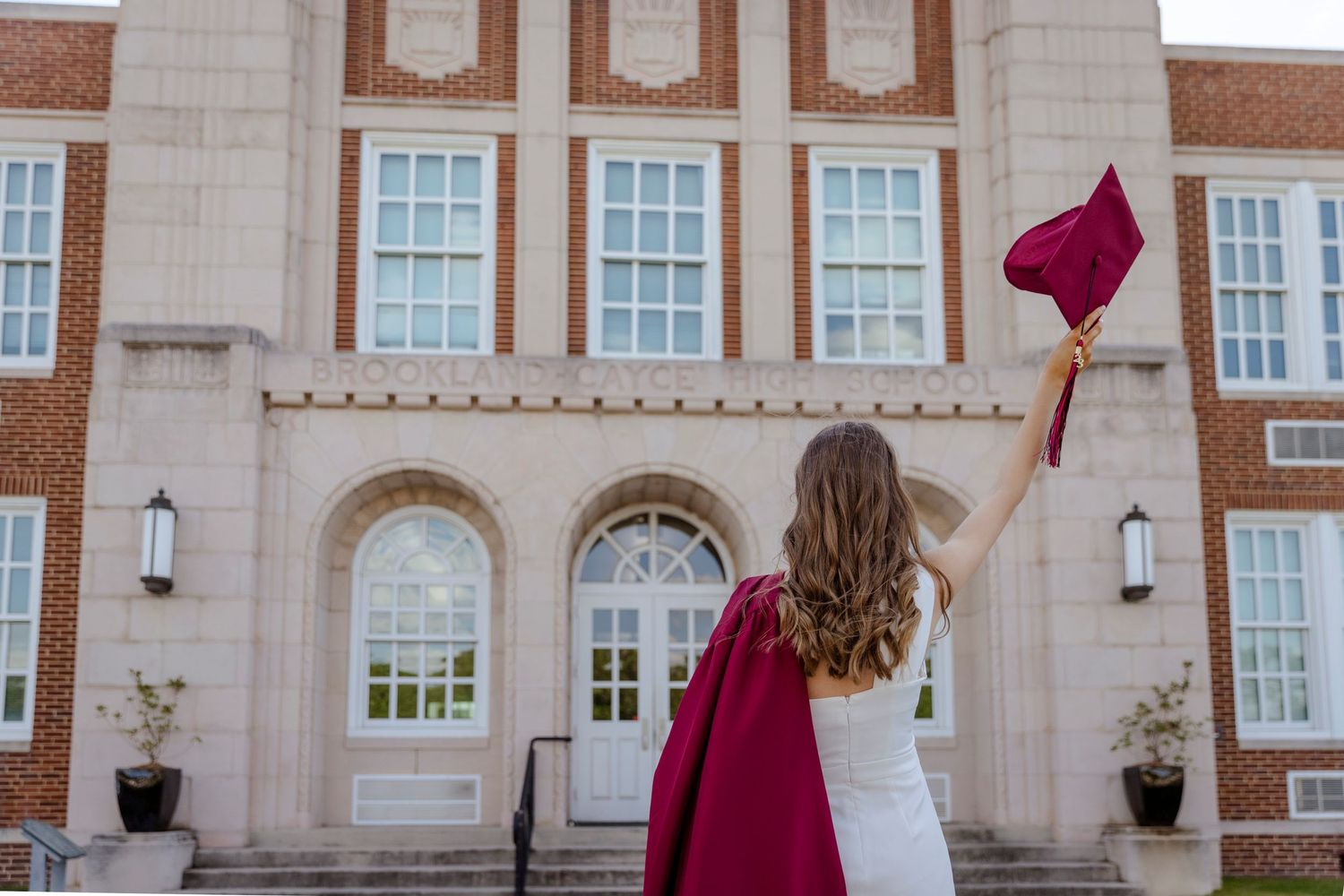 A graduate in white and red holds up their graduation cap in front of a brick high school building.