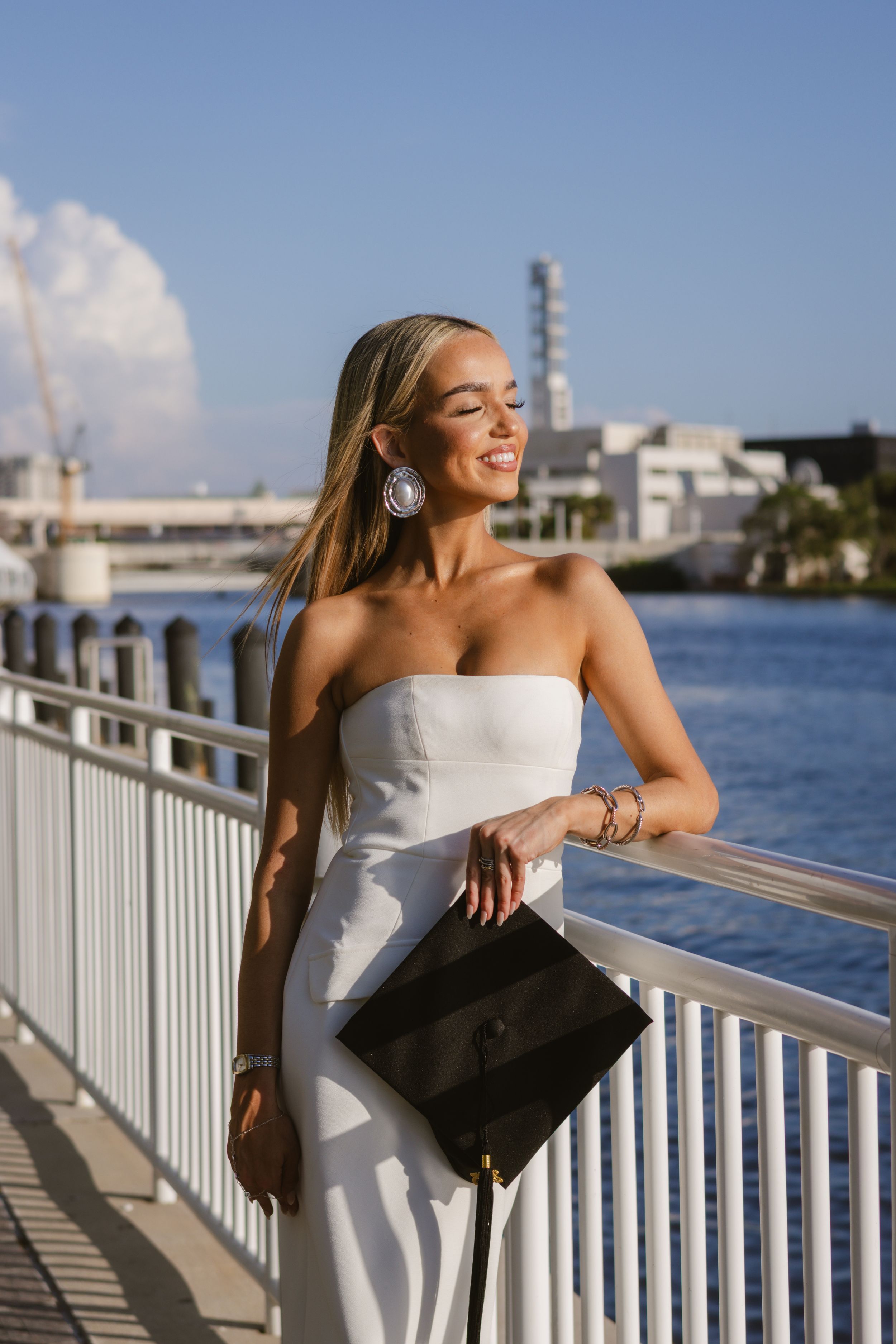 A girl in a white dress is leaned on the railing at Curtis Hixon park. She is holding a graduation cap, and looking toward the water & sun.