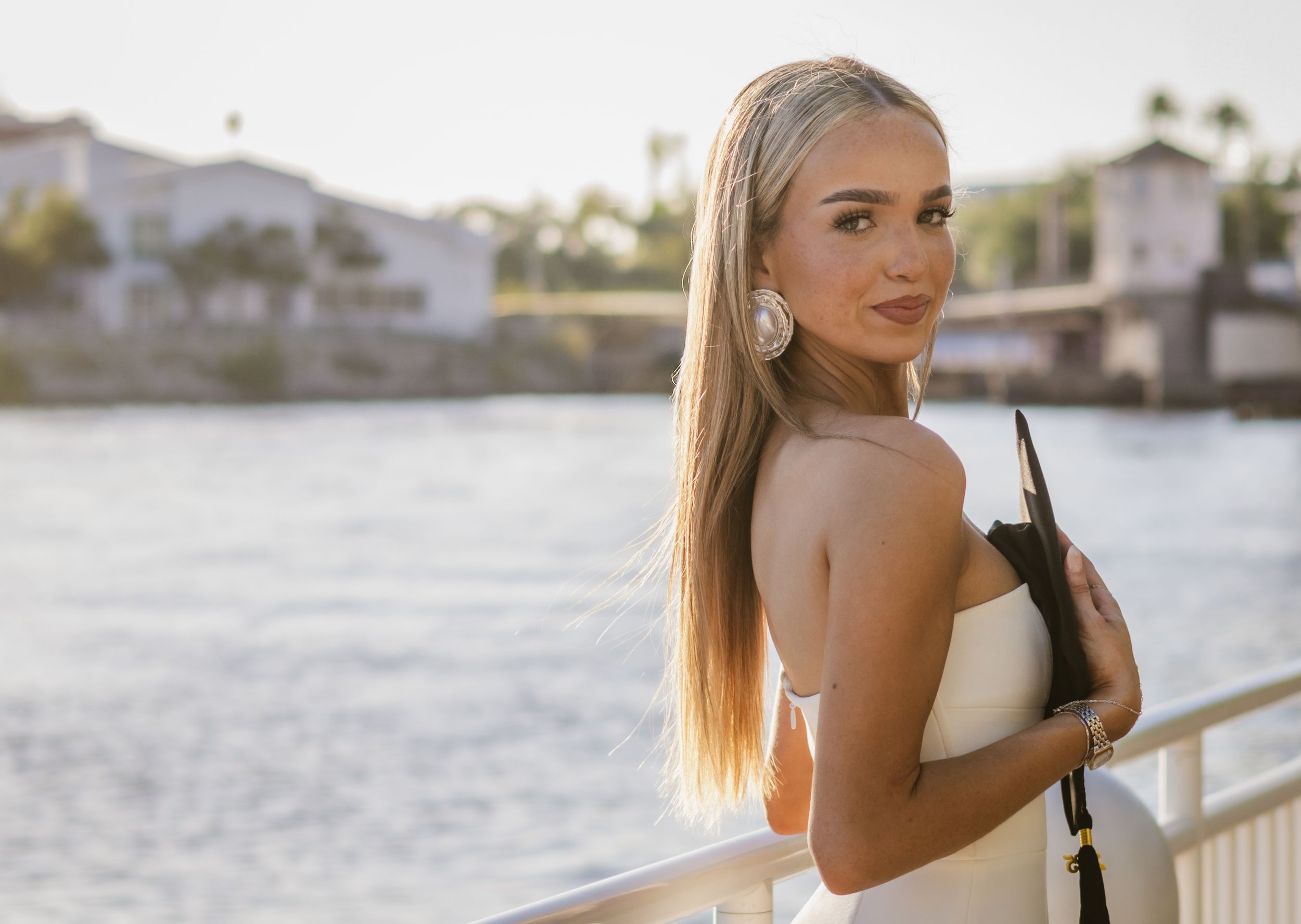Hugging her graduation cap to her chest, a girl looks over her shoulder, as the Tampa river glows behind her. At Curtis Hixon Park.