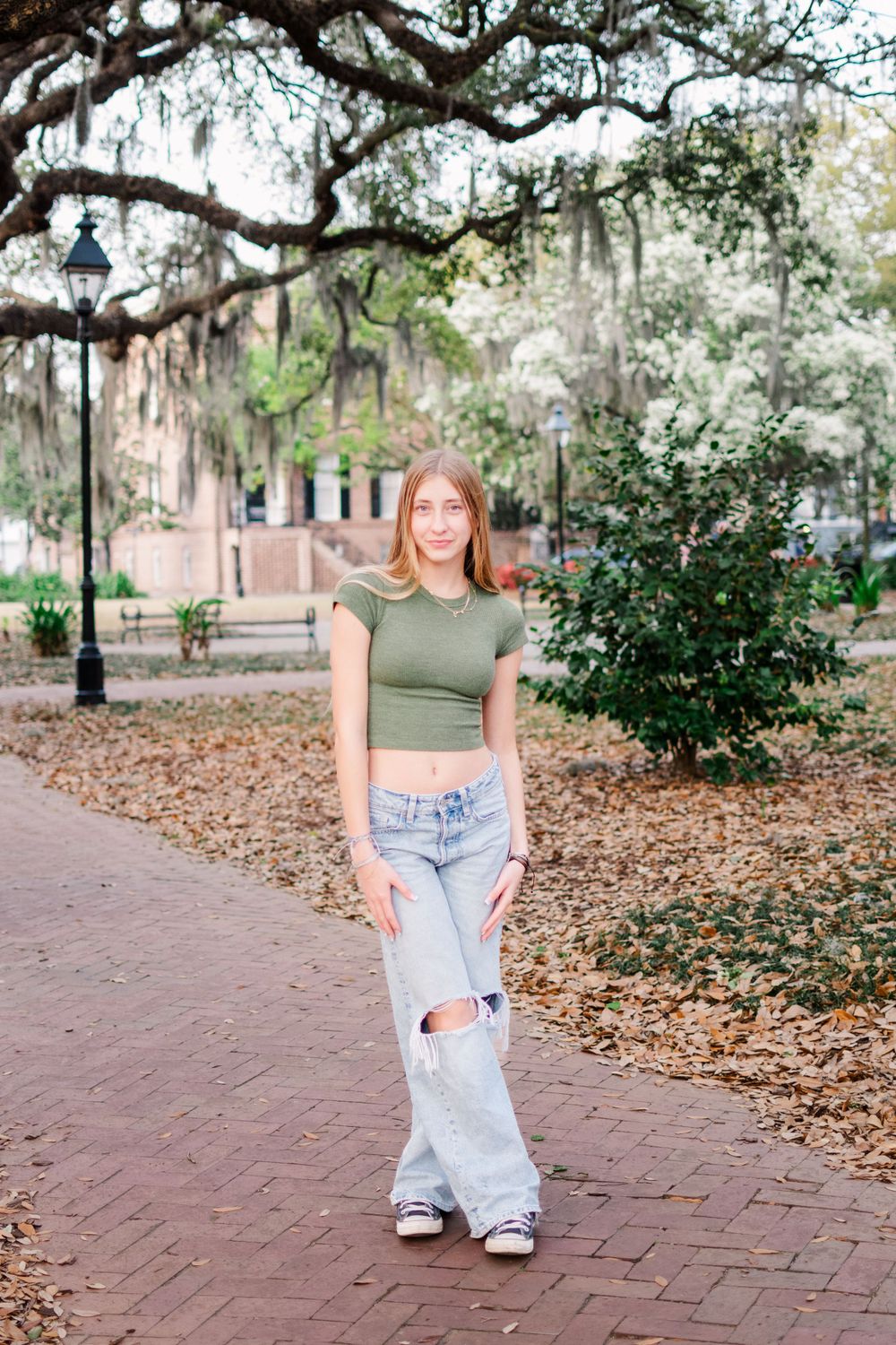Person in green crop top and ripped jeans poses on a brick path under Spanish moss trees.