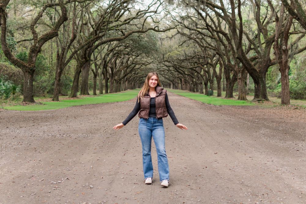 Person poses with arms outstretched on dirt road under canopy of oak trees draped with Spanish moss.