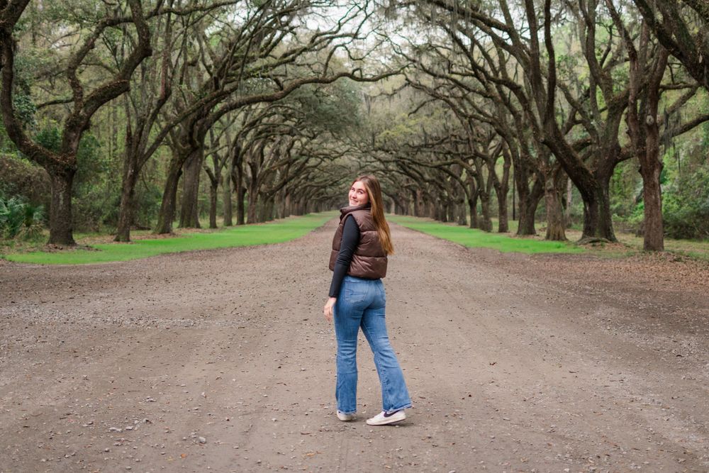 Person standing on a dirt road under a canopy of Spanish moss-covered oak trees in a scenic park.