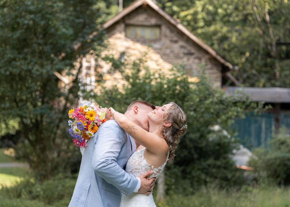 Lachende Braut mit buntem Wildblumenstrauß in den Armen ihres Bräutigams vor altem Steinhaus, Sommerhochzeit bei Karlsruhe