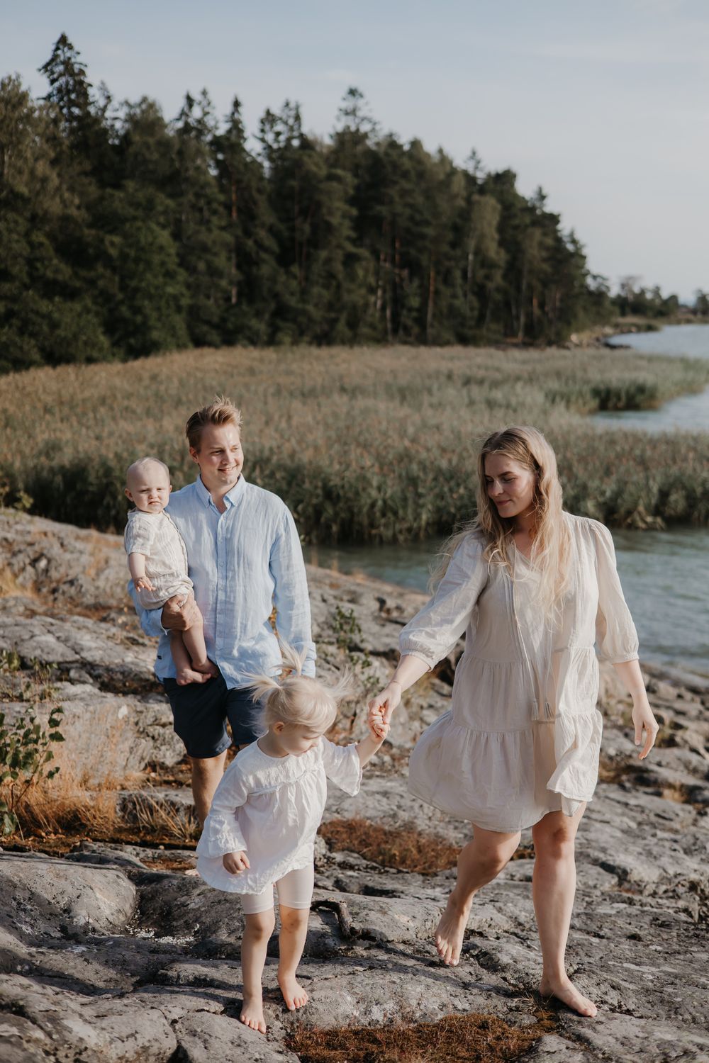 A family explores rocky coastal terrain surrounded by evergreen trees and marsh grasses at sunset.