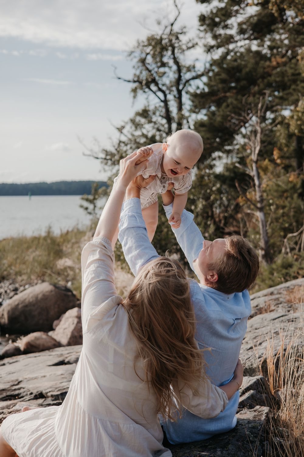 A couple lifts their baby high in the air while sitting on coastal rocks near water.