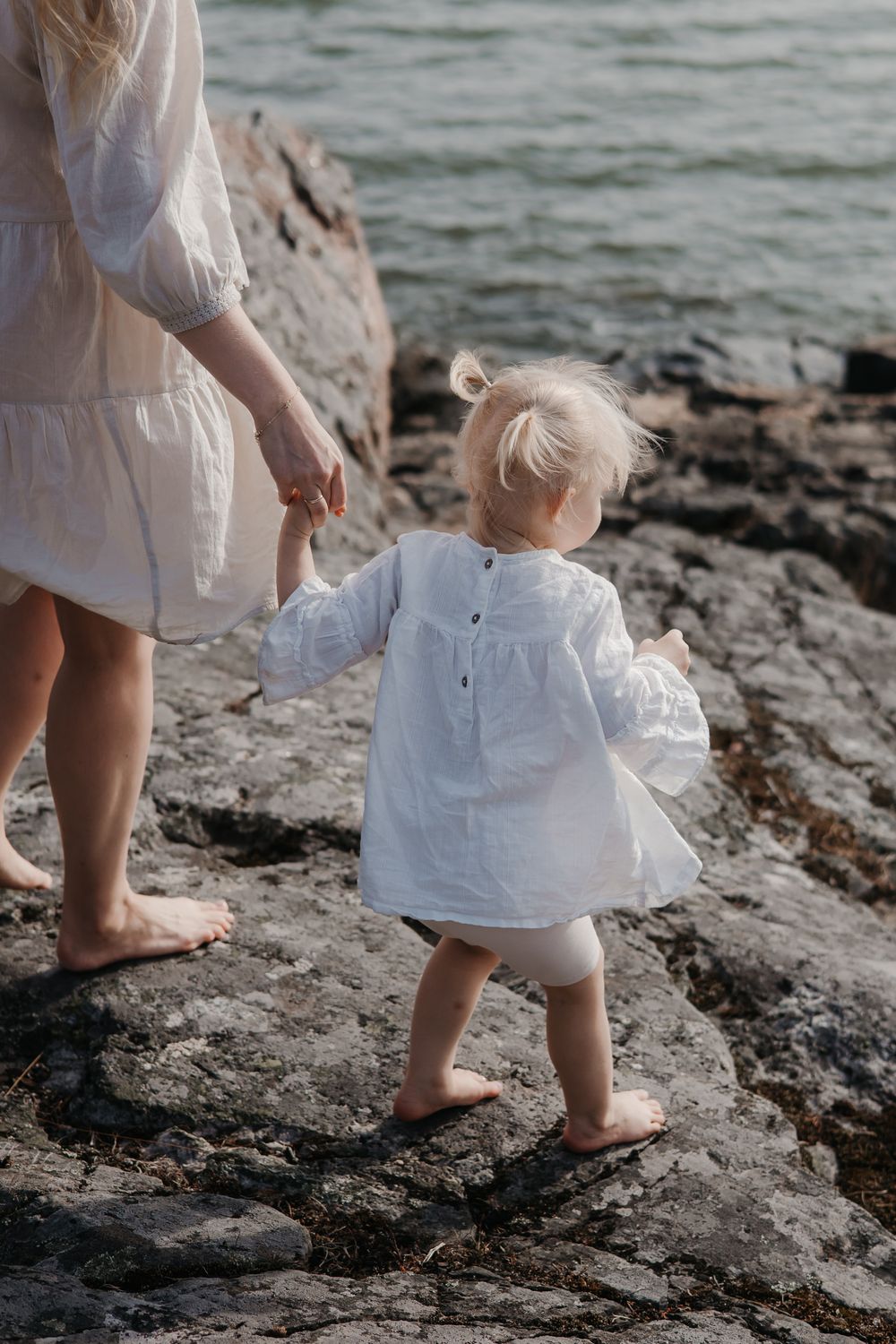 A toddler in a white dress holds hands while walking on rocky shoreline.