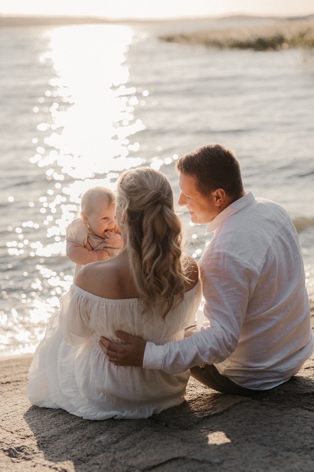 A young family sits by a sunlit shore, parents gazing lovingly at their baby in a golden sunset.