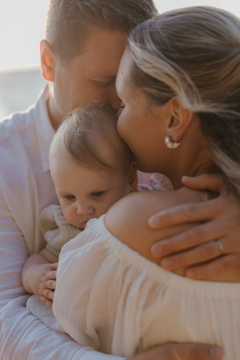 A couple tenderly kisses their baby during a soft, golden-hour family portrait session outdoors.
