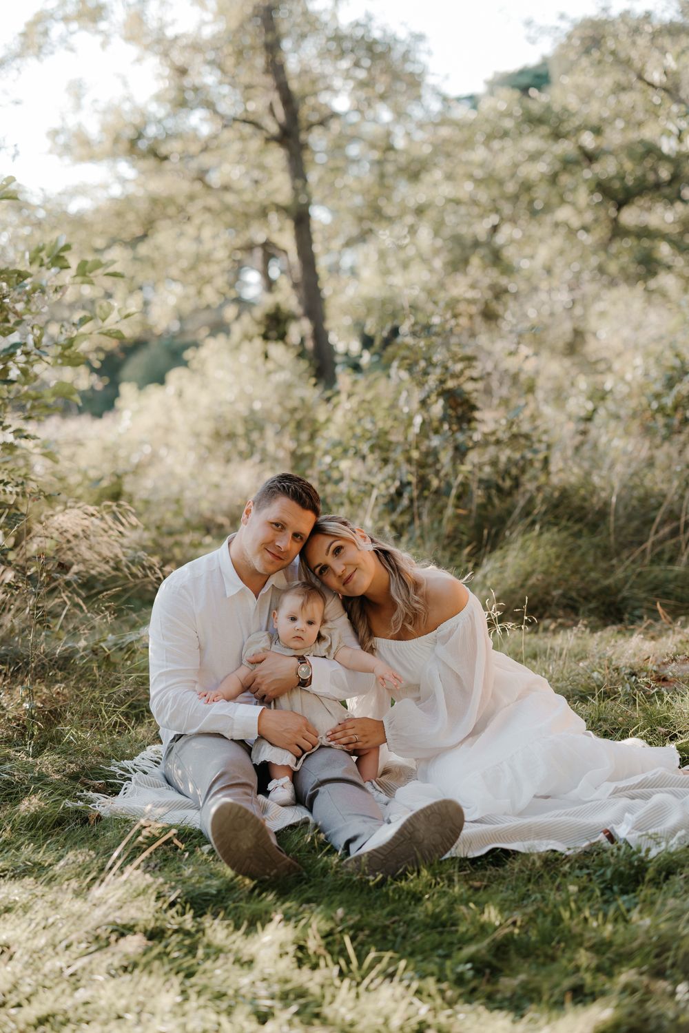 A young family sits on a white blanket in a sunny meadow, parents in white clothing cradling their baby outdoors.