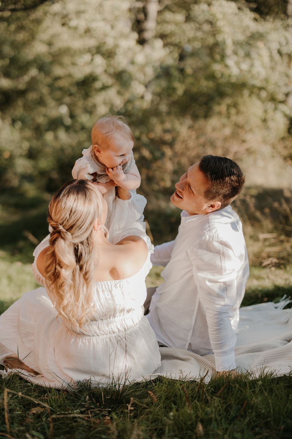 A young family in white outfits sits on a blanket outdoors, with parents smiling at their happy baby.