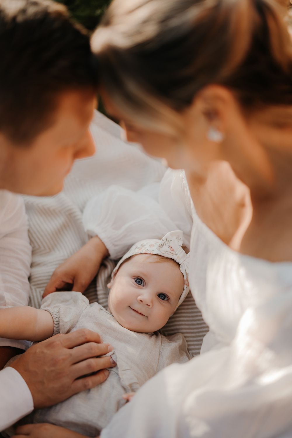A smiling baby in a white outfit and headband looks up at two adults in white clothing during a family photo session.