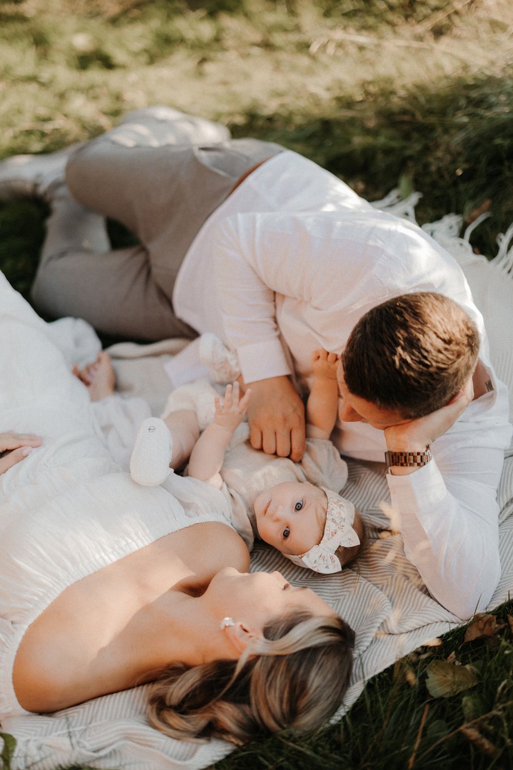 A couple lies on a white blanket outdoors, lovingly gazing at their baby girl in a heartwarming family moment.