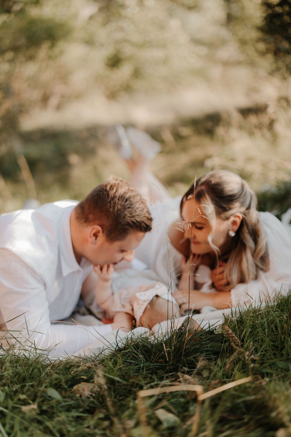 A couple lies on green grass, gazing lovingly at their newborn baby in a serene outdoor setting.