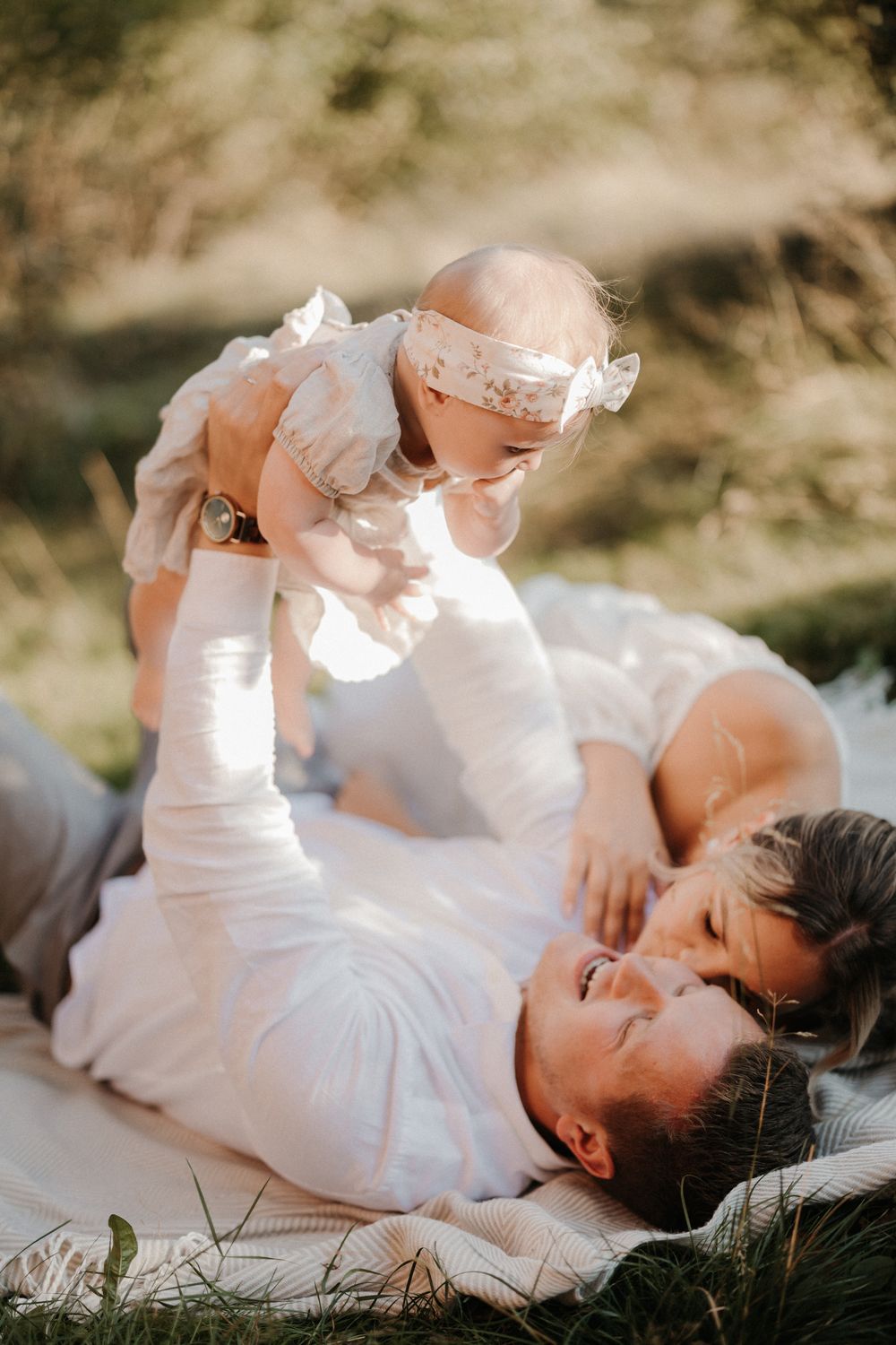 Parents lying on a blanket outdoors, lovingly lifting their baby girl in a white dress in warm sunlight.