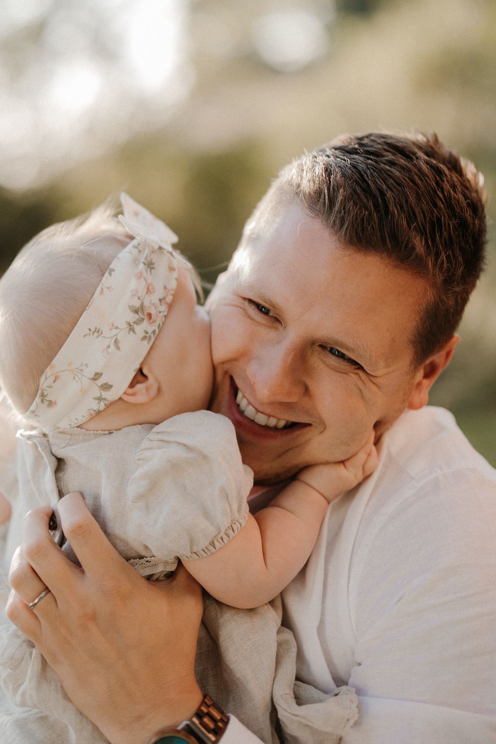 Smiling father holding baby girl in floral headband, sharing a tender cheek kiss outdoors.