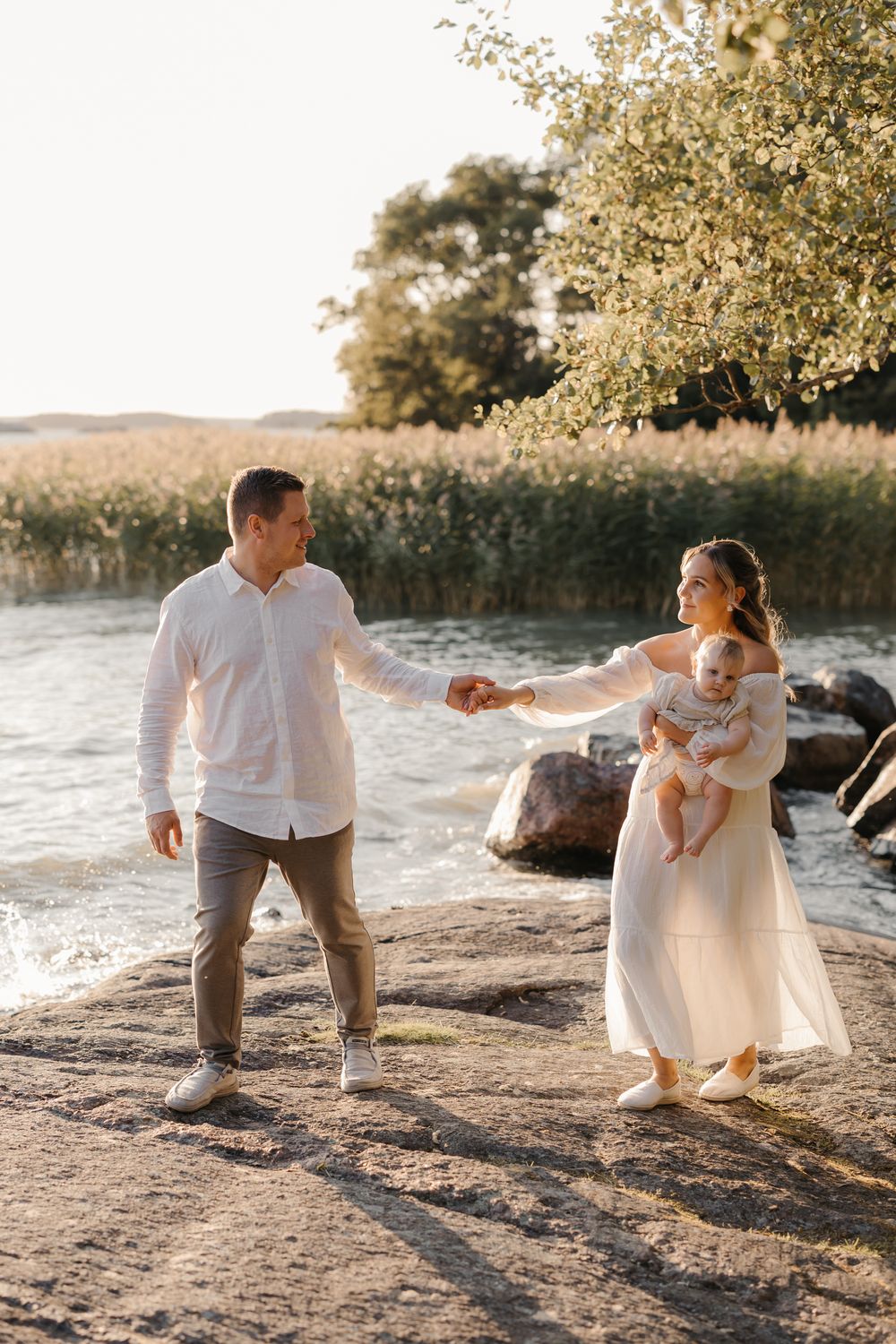 A couple holding hands near a lake, with the woman carrying a baby, dressed in white, during golden hour.