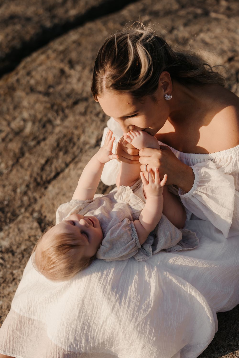 A mother in a white dress tenderly kisses her baby's feet while the infant lies in her lap outdoors.
