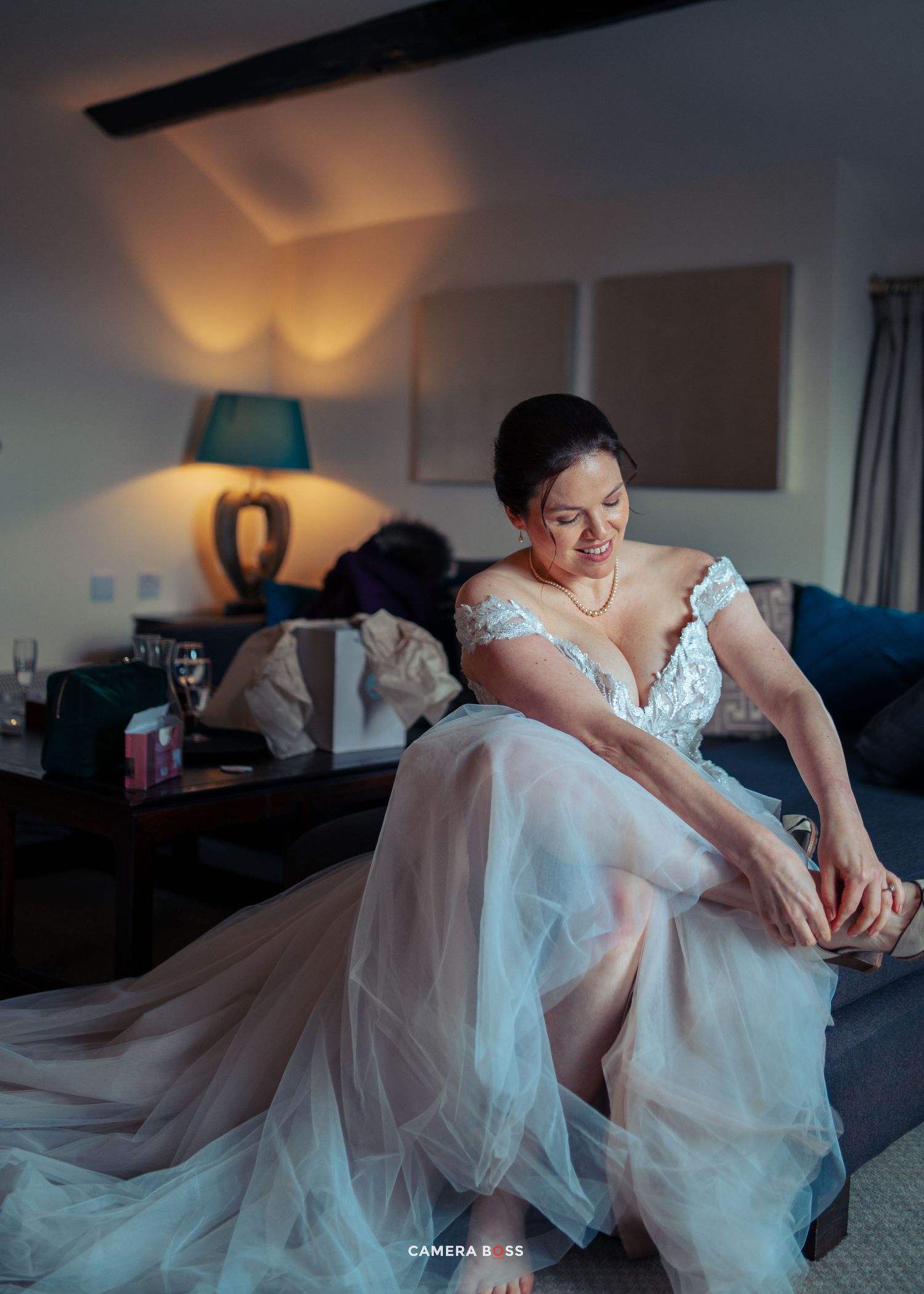 A bride in a flowing white lace gown sits on a couch while adjusting her shoe in warm ambient lighting.