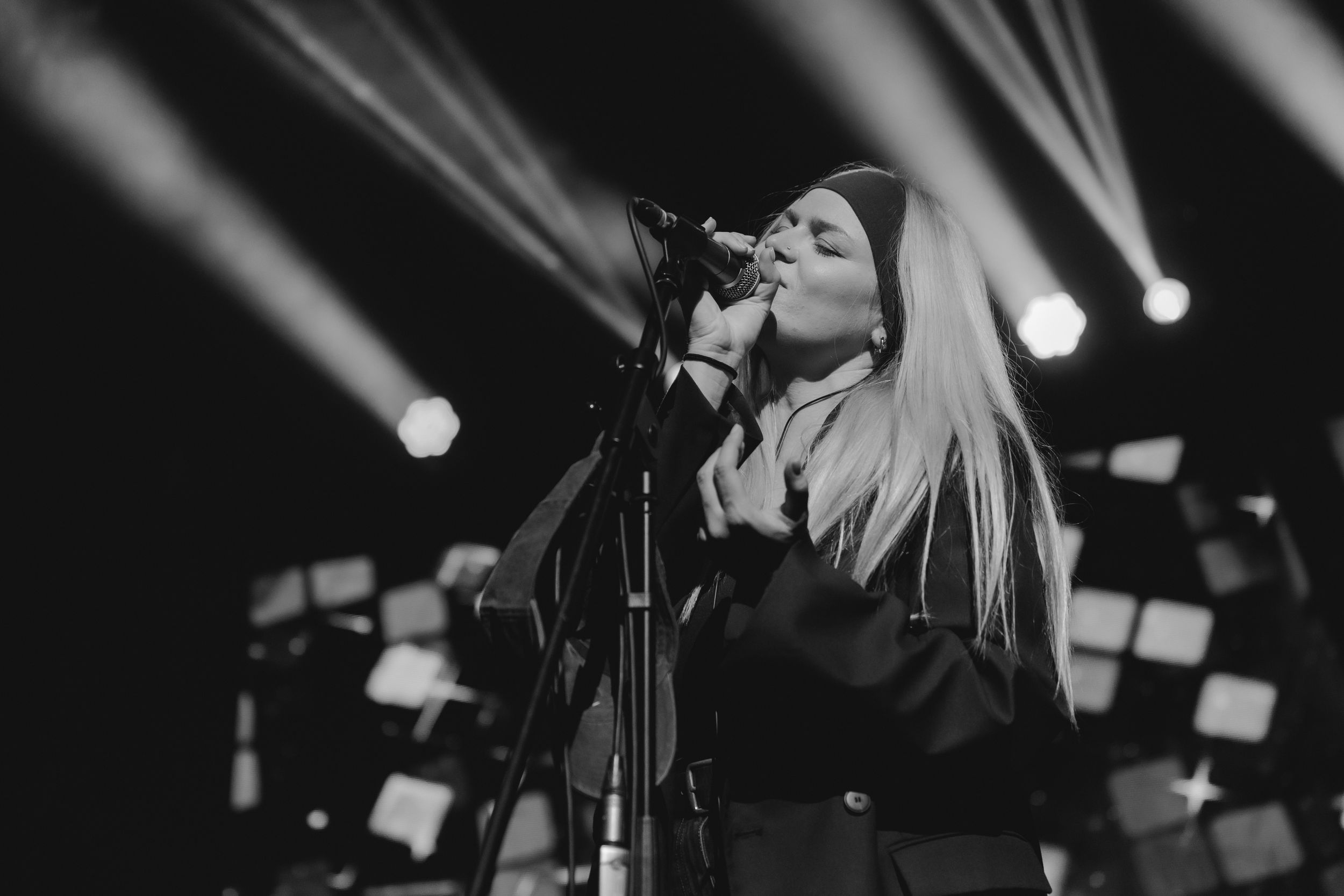 Singer performs passionately on stage with microphone in black and white photo with dramatic concert lighting in background.