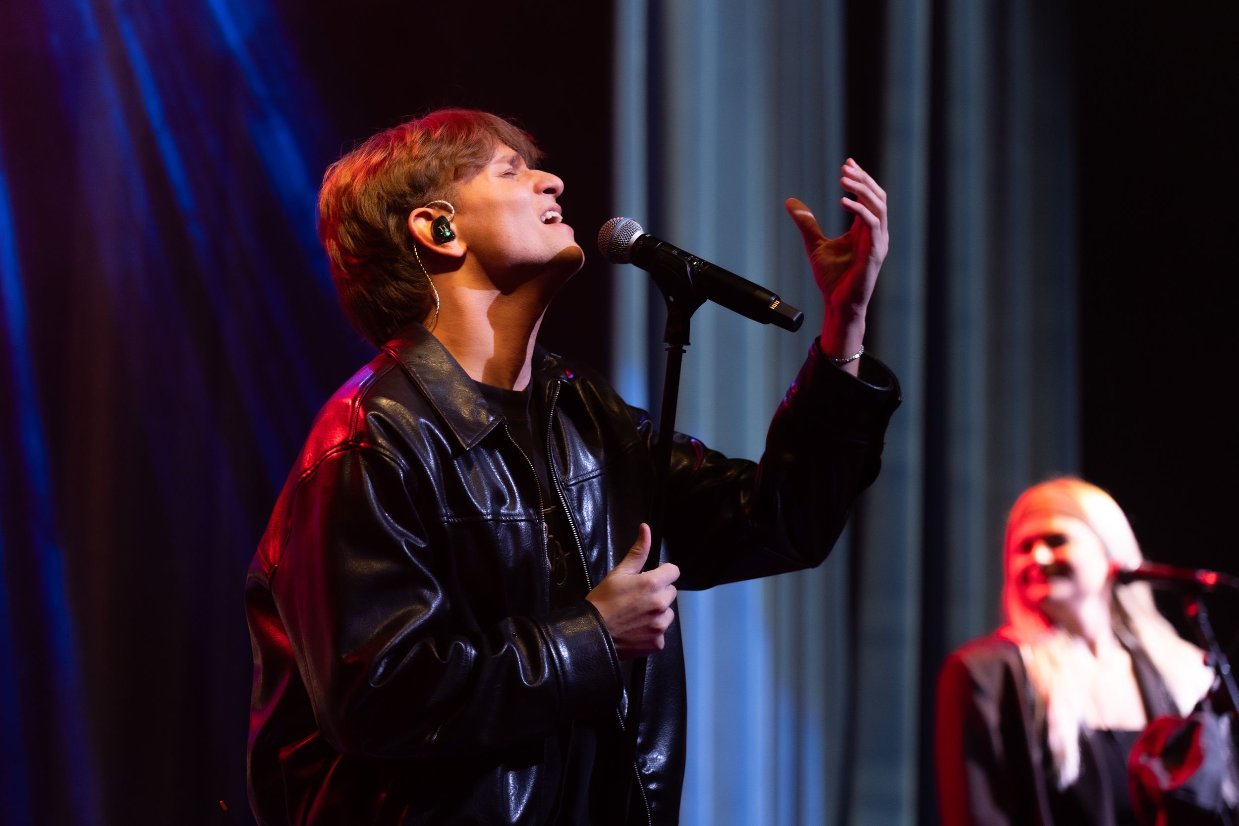 A performer sings passionately into a microphone on stage under dramatic blue and red lighting.
