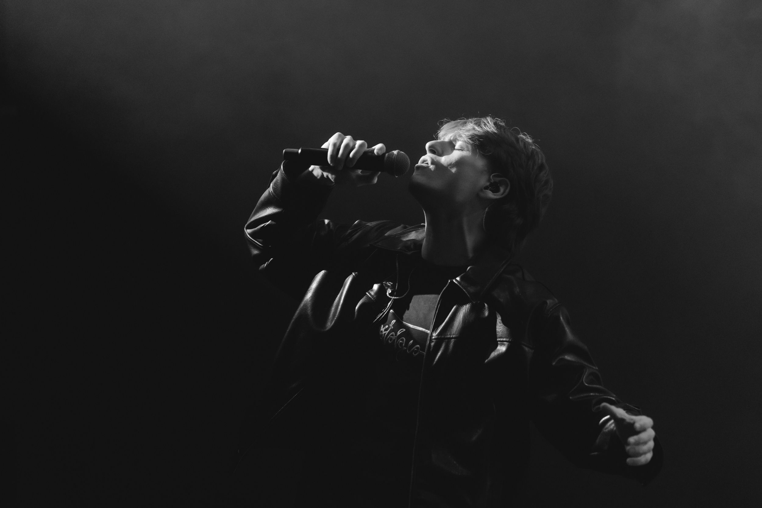 A dramatic black and white performance photo of a singer silhouetted against darkness while singing into a microphone.