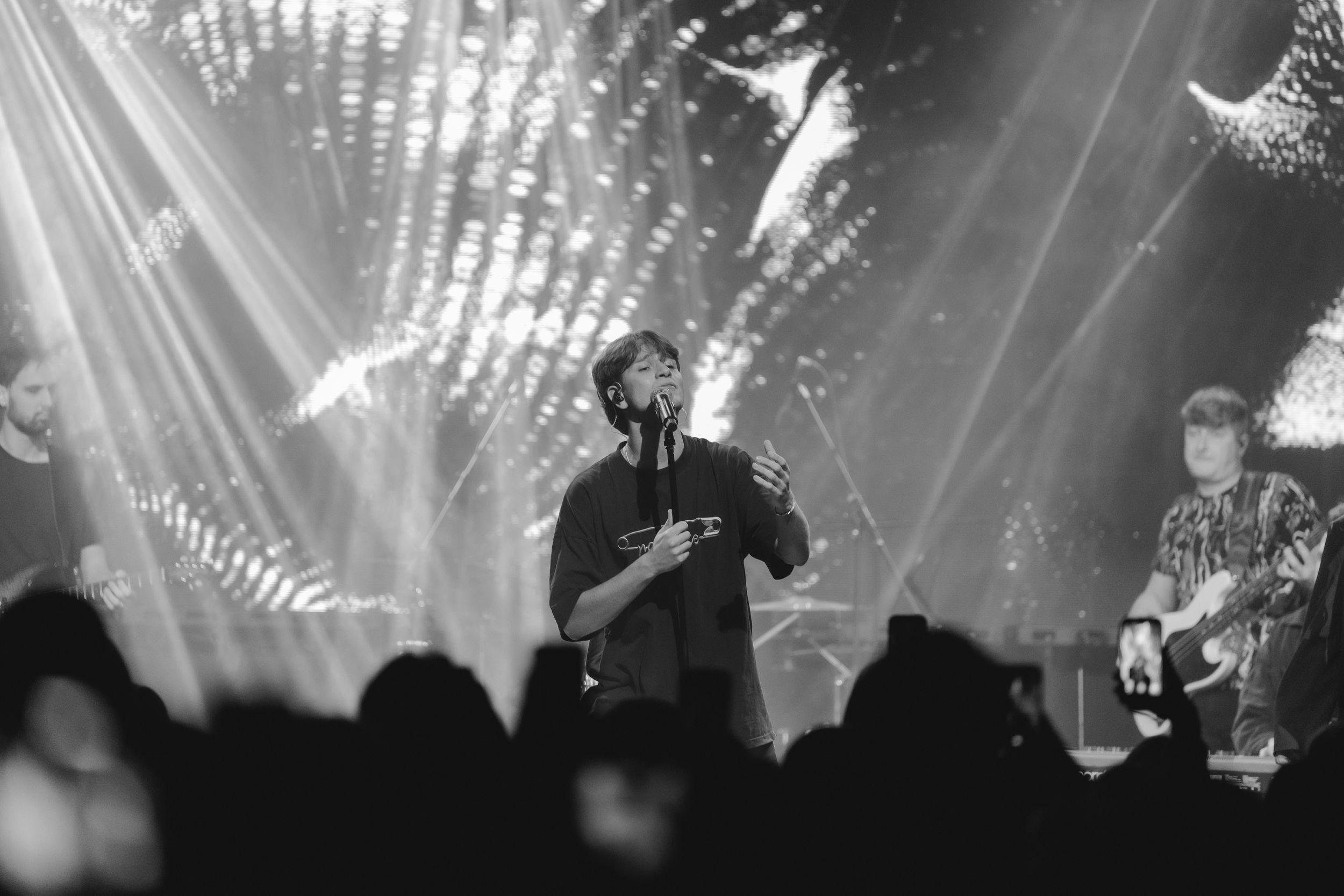 Black and white concert photo showing a performer on stage with dramatic lighting beams and silhouetted crowd in foreground.