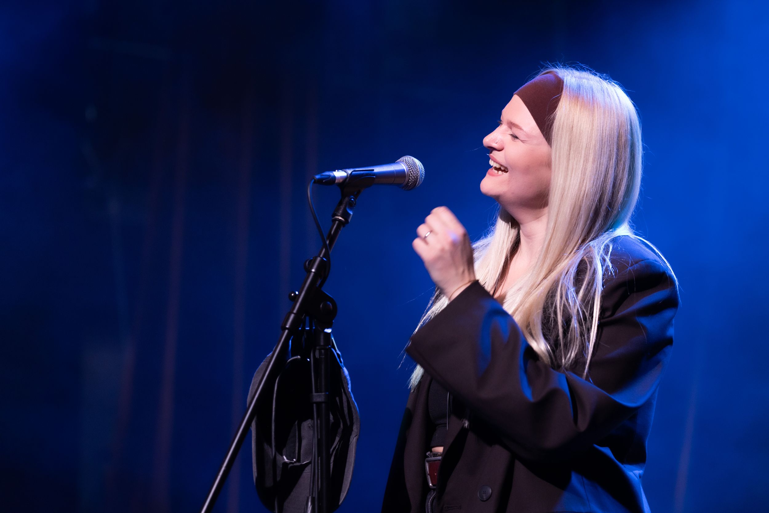 A performer in a black blazer sings passionately into a microphone on stage with dramatic blue lighting.