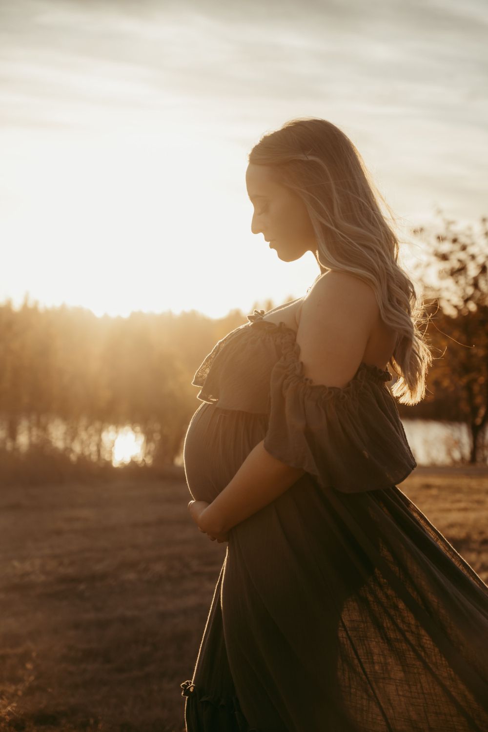 Silhouette of a pregnant woman in flowing dress standing against golden sunset by a lake.